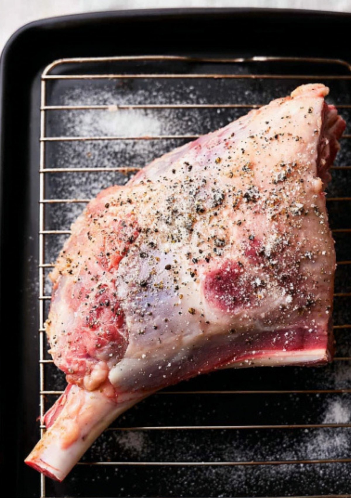 Raw bone-in leg of lamb seasoned with salt and black pepper on a rack inside a roasting pan