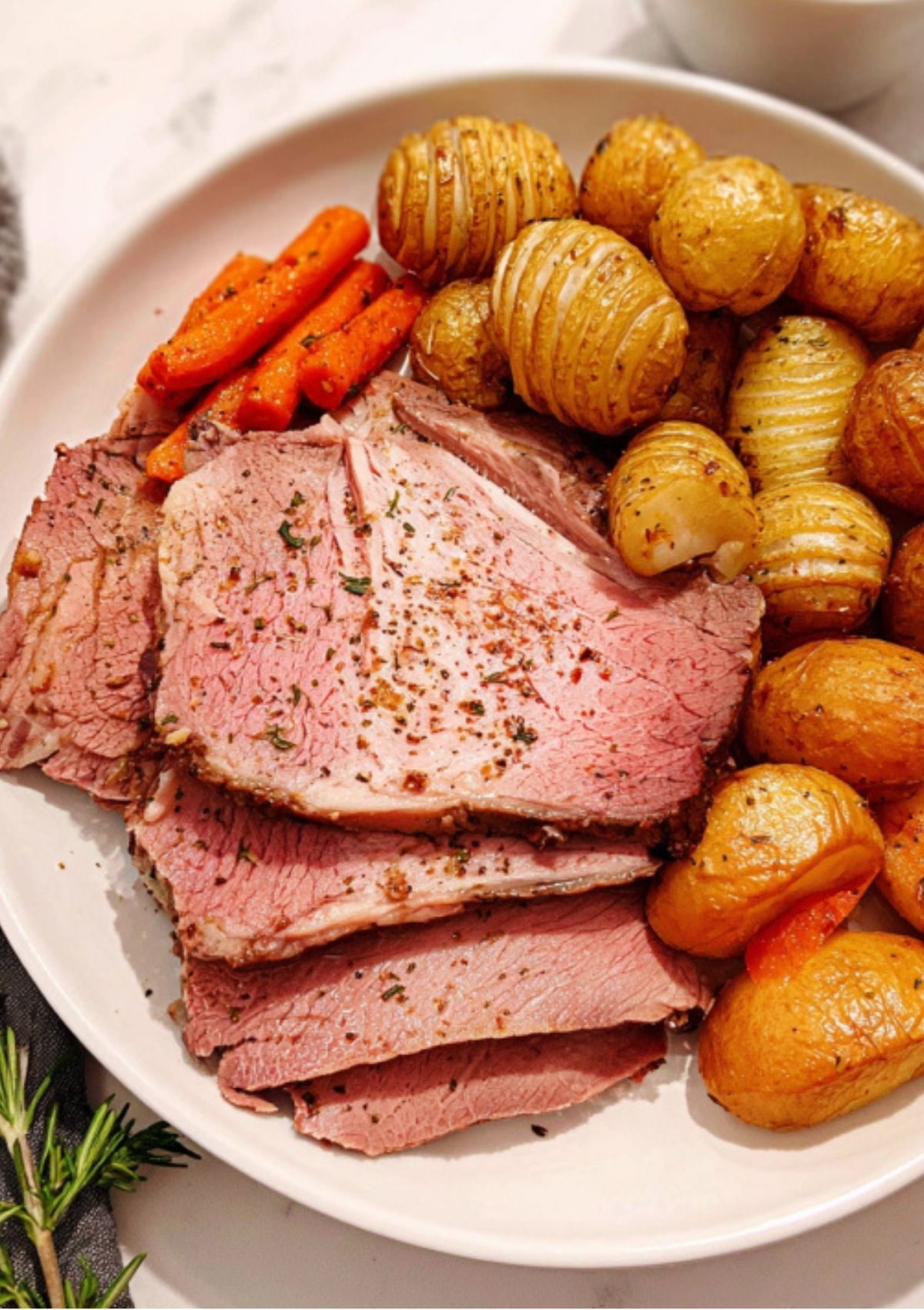 Slices of juicy oven roasted leg of lamb served with crispy hasselback potatoes and roasted carrots on a white plate.