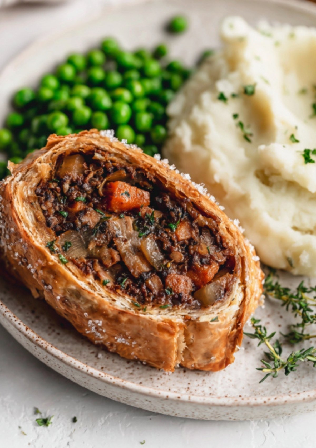 Close up of a crispy vegan Wellington filled with plant-based ground, carrots, peas, and mushrooms, paired with classic mashed potatoes and peas. Great centerpiece for vegan holiday feasts.