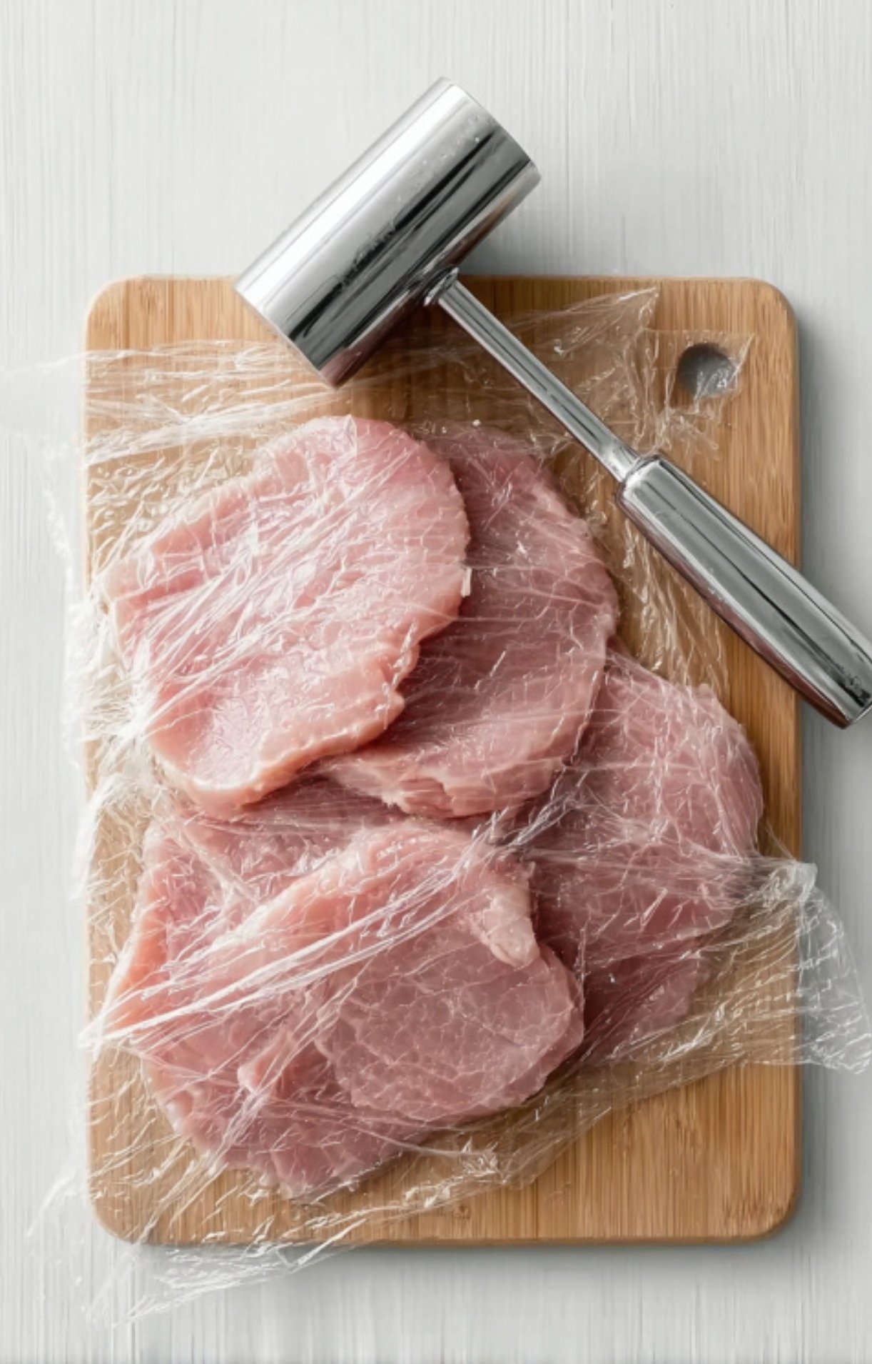 Raw veal cutlets covered with plastic wrap and flattened with a metal meat mallet on a wooden cutting board, ready to be used for veal scallopini.
