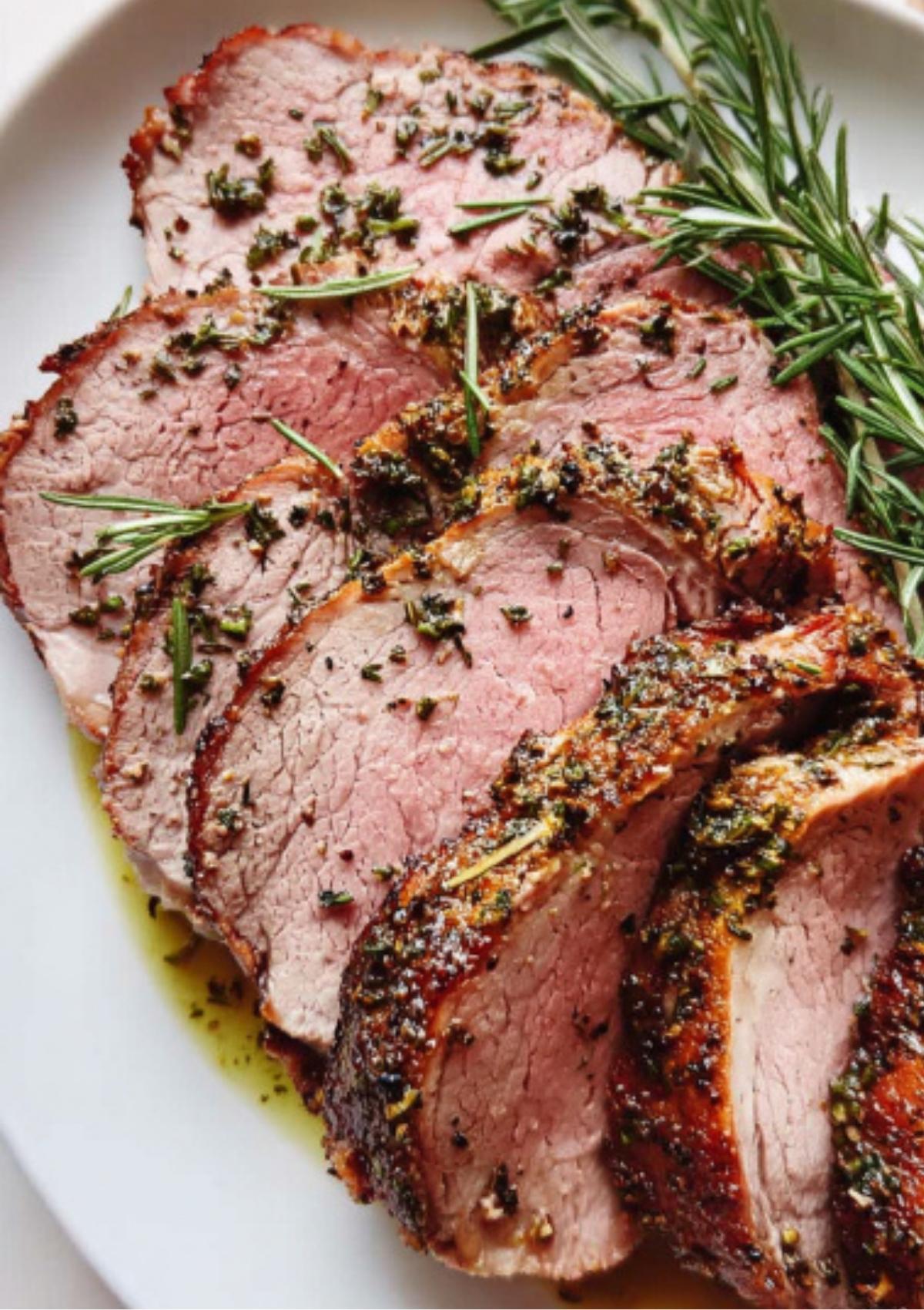 Slices of roasted leg of lamb with a garlic rosemary crust arranged on a white plate with fresh rosemary