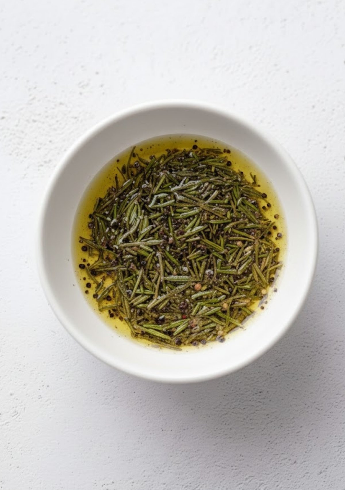 White bowl filled with olive oil, rosemary, cracked pepper, and sea salt for lamb marinade
