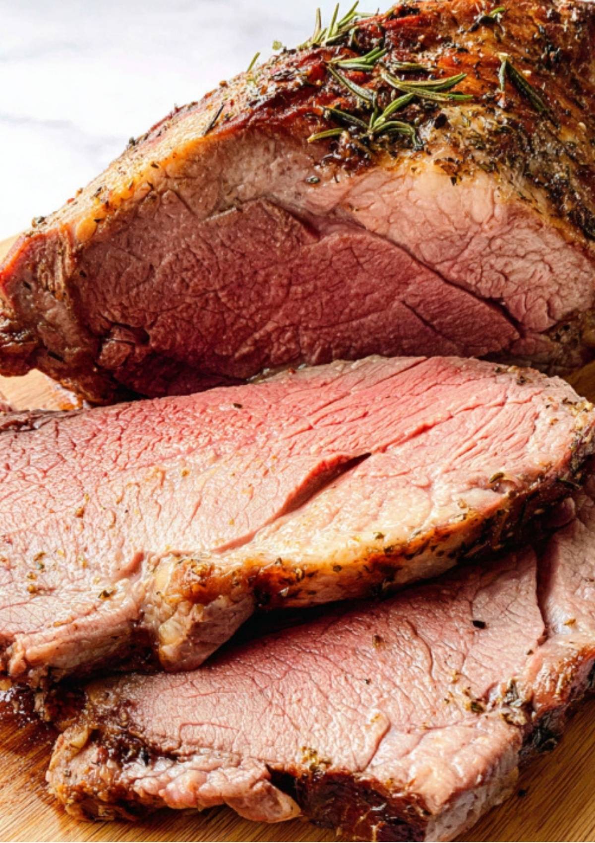 Slices of tender, medium-rare roasted leg of lamb with a golden crust, garnished with rosemary.