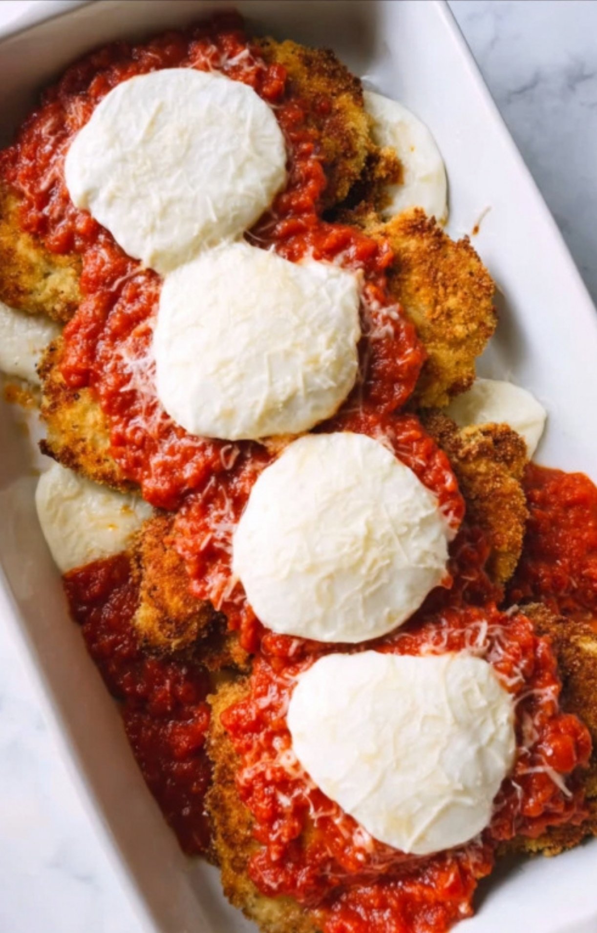 Breaded veal in a baking dish topped with red sauce and rounds of mozzarella, ready for baking