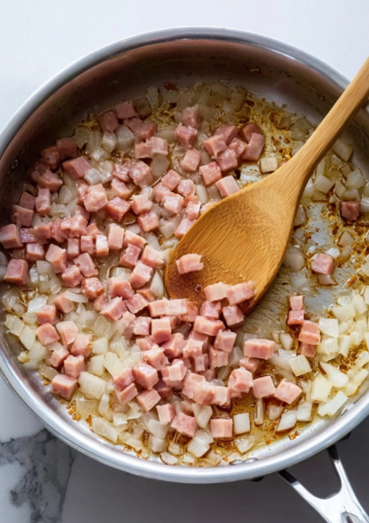 Diced ham and onions being sautéed in a stainless steel skillet, stirred with a wooden spoon.