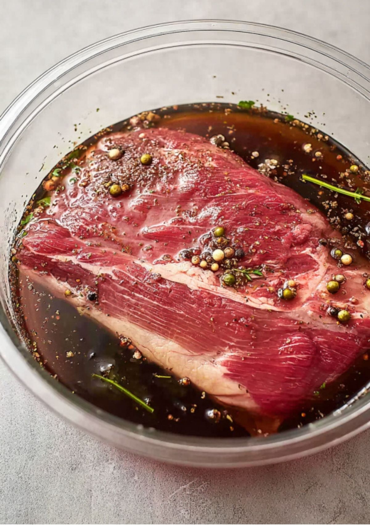Uncooked brisket submerged in a clear bowl of seasoned brine with mustard seeds, peppercorns, and fresh herbs, starting the curing process.