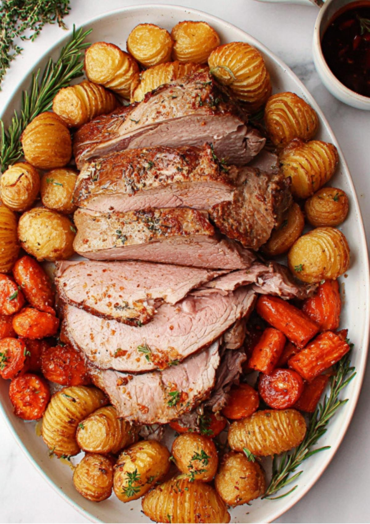 Platter of oven roasted leg of lamb sliced and served with crispy hasselback potatoes, roasted carrots, and fresh herbs.