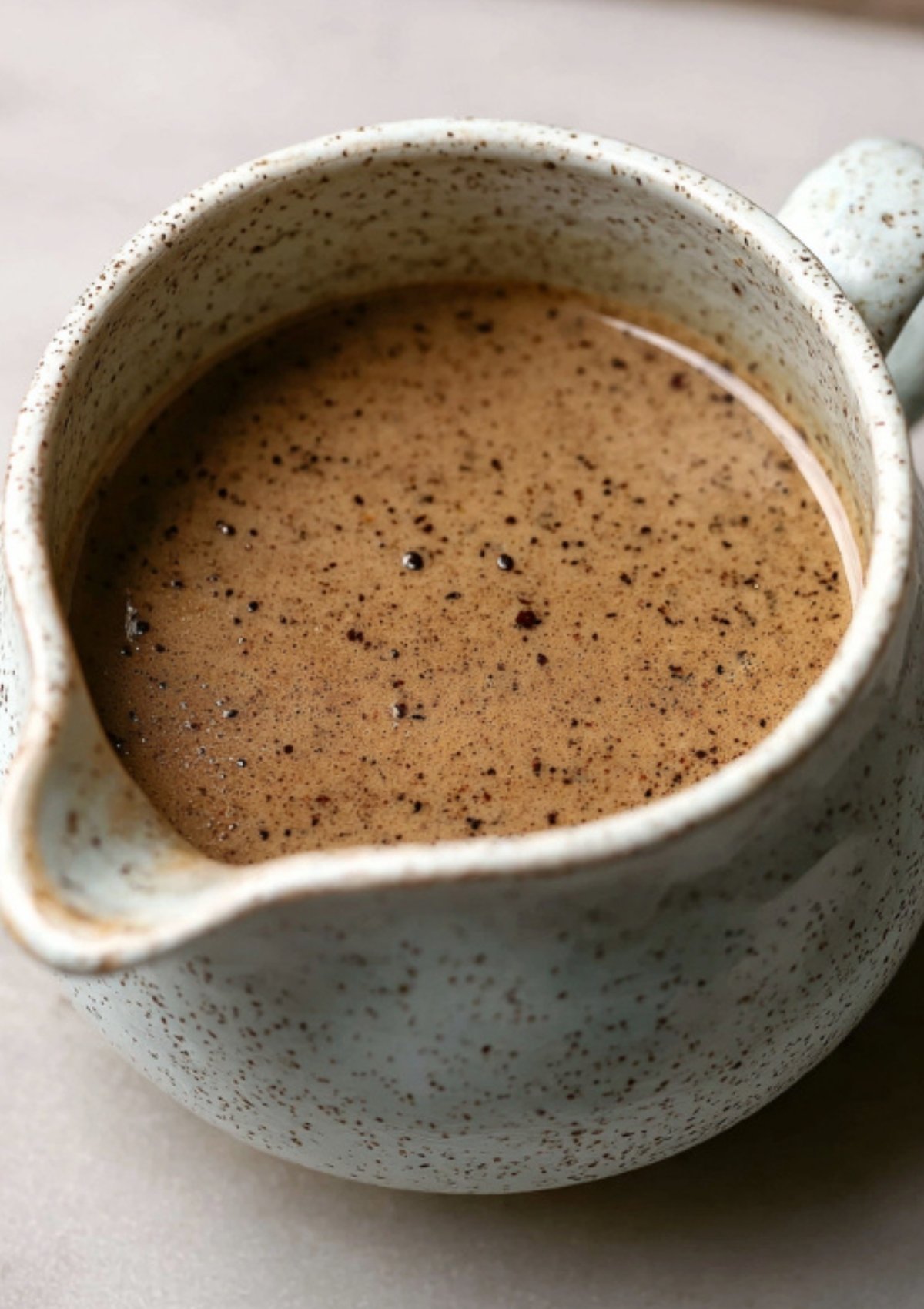 Closeup of homemade creamy black peppercorn sauce in a rustic speckled jug, perfect for serving with Beef Wellington, steak, or festive roast dinners.