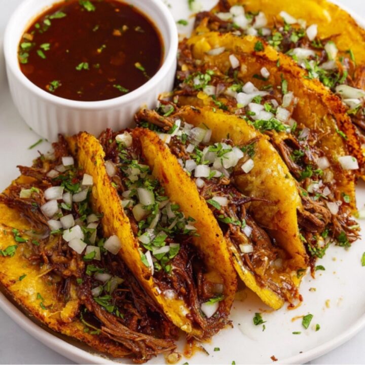A plate of four crispy beef birria tacos topped with white onion and cilantro, served with a small bowl of rich red dipping broth.