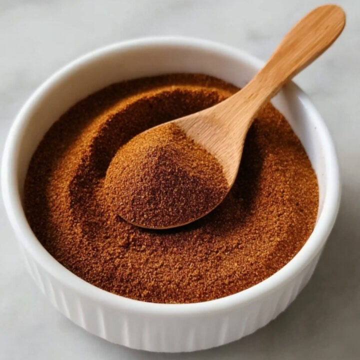 A close-up shot of a wooden spoon lifting a portion of homemade Birria Taco Seasoning from a white bowl, highlighting the rich color and earthy texture.