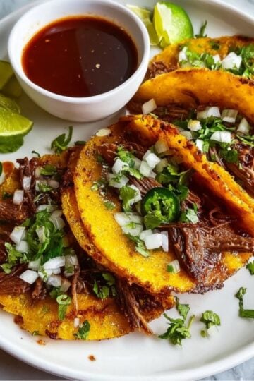 A white plate featuring three golden-brown birria tacos garnished with cilantro and onions, served with lime, jalapeños, and a side of red consommé.