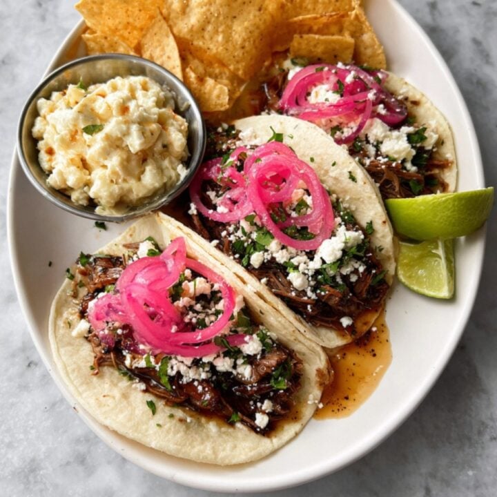 Three fully assembled chuck roast tacos served on a white plate with tortilla chips, a side of corn salad, and fresh lime wedges.