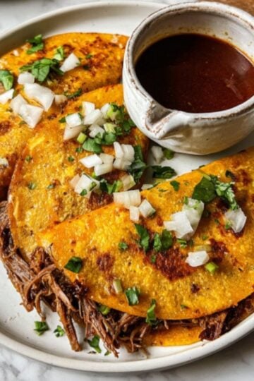 A professional food photo of crispy, golden-brown Crockpot Birria Tacos garnished with fresh cilantro and onions next to a bowl of consommé.