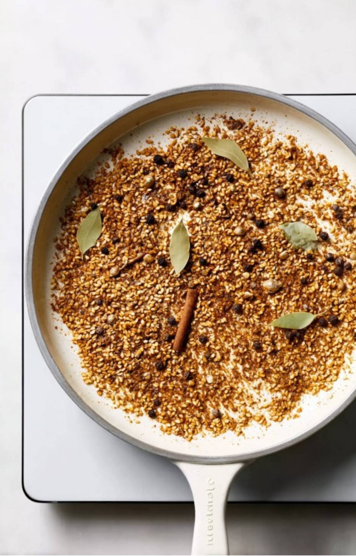 A white pan filled with toasted sesame seeds, black peppercorns, bay leaves, and a cinnamon stick being dry-roasted.