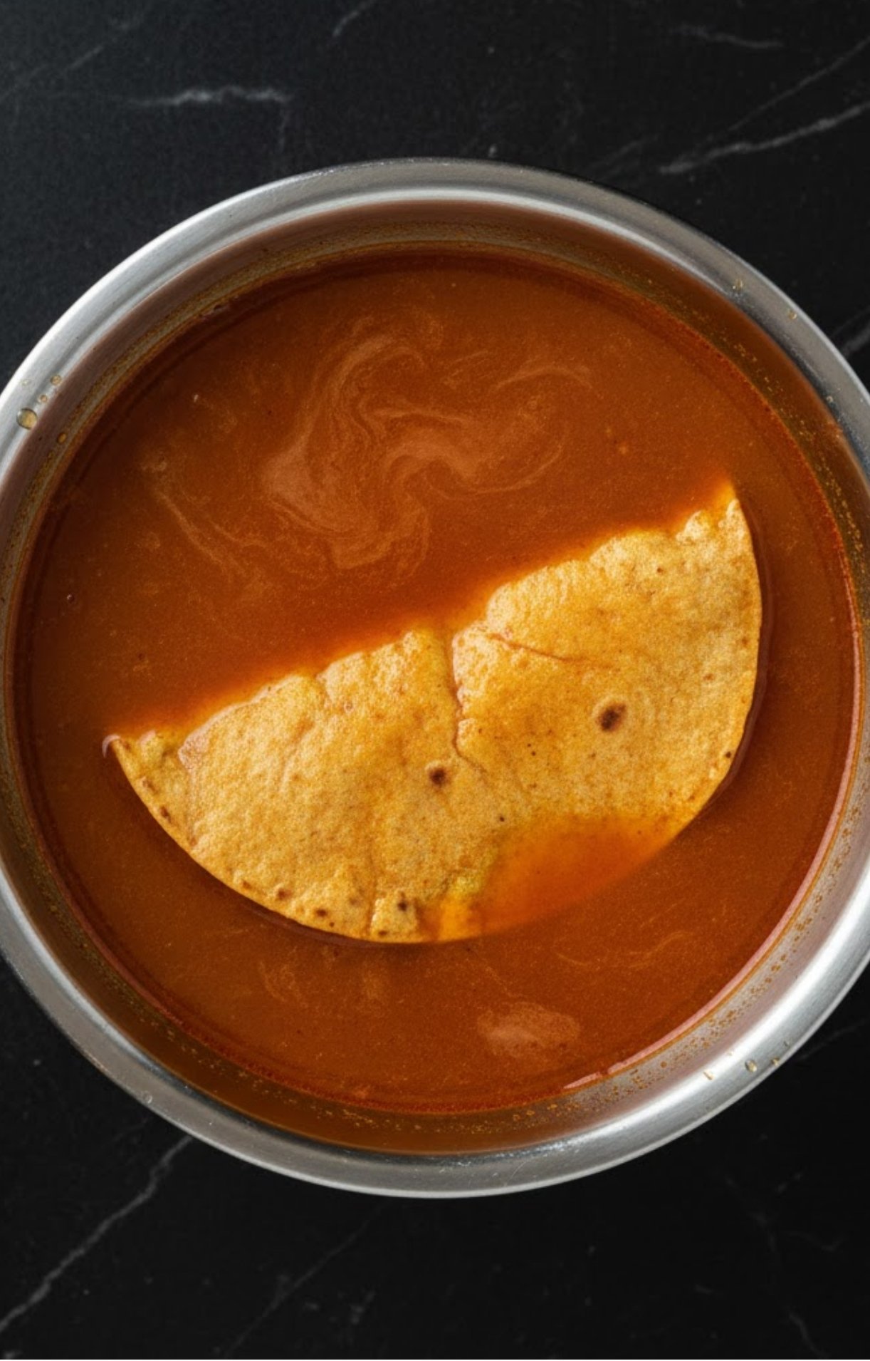 A yellow corn tortilla being dipped into a stainless steel bowl filled with rich, reddish-brown birria consommé to coat it in fat before frying.