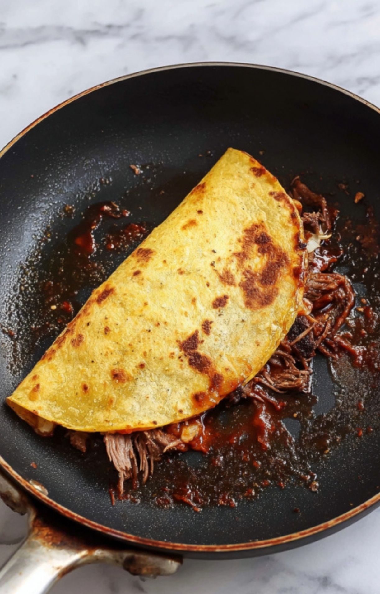 A single corn tortilla folded over shredded beef and cheese, frying in a pan with red birria oil to create a crispy shell.
