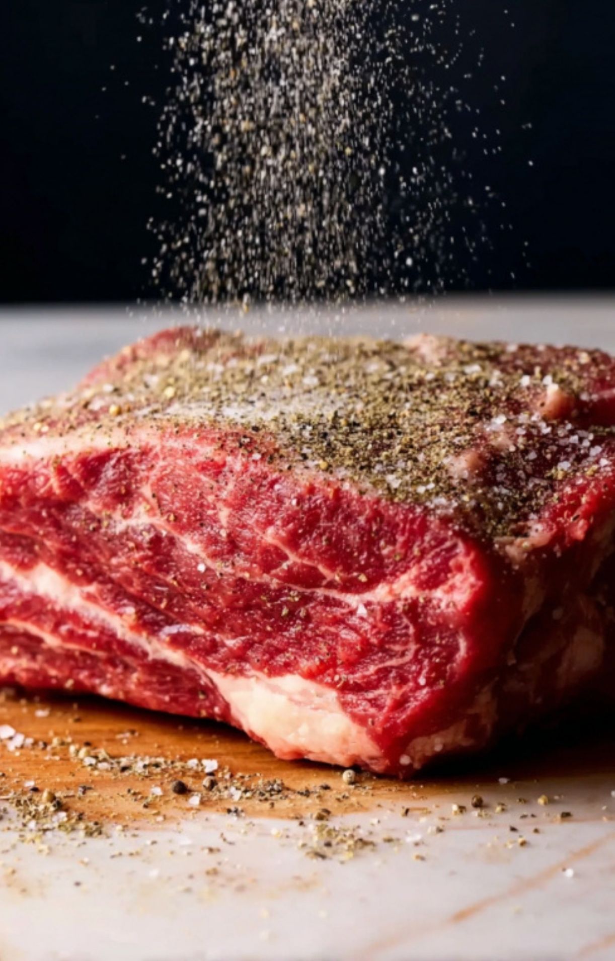 Hand sprinkling a generous layer of salt and coarse black pepper onto a thick, marbled raw chuck roast.