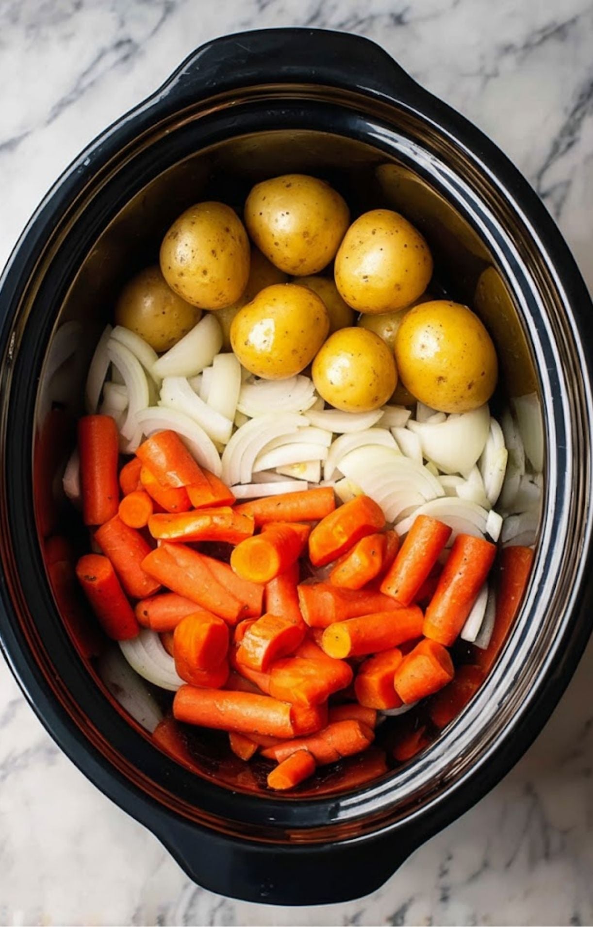 The base of a slow cooker filled with a layer of sliced white onions, topped with baby carrots and whole yellow potatoes.