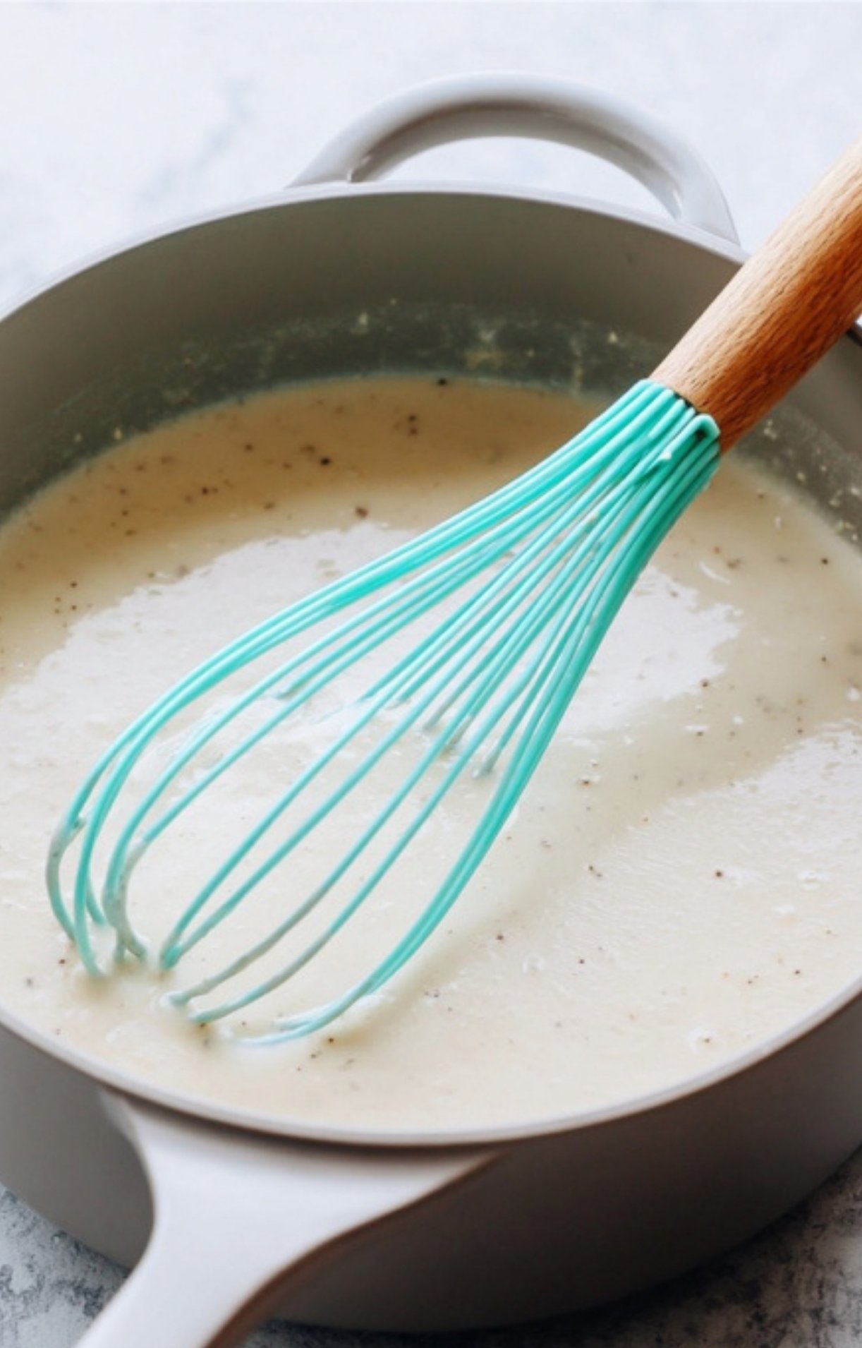 A turquoise whisk stirring a creamy white sauce with black pepper in a pot to create the base for Easy Turkey Pot Pie.