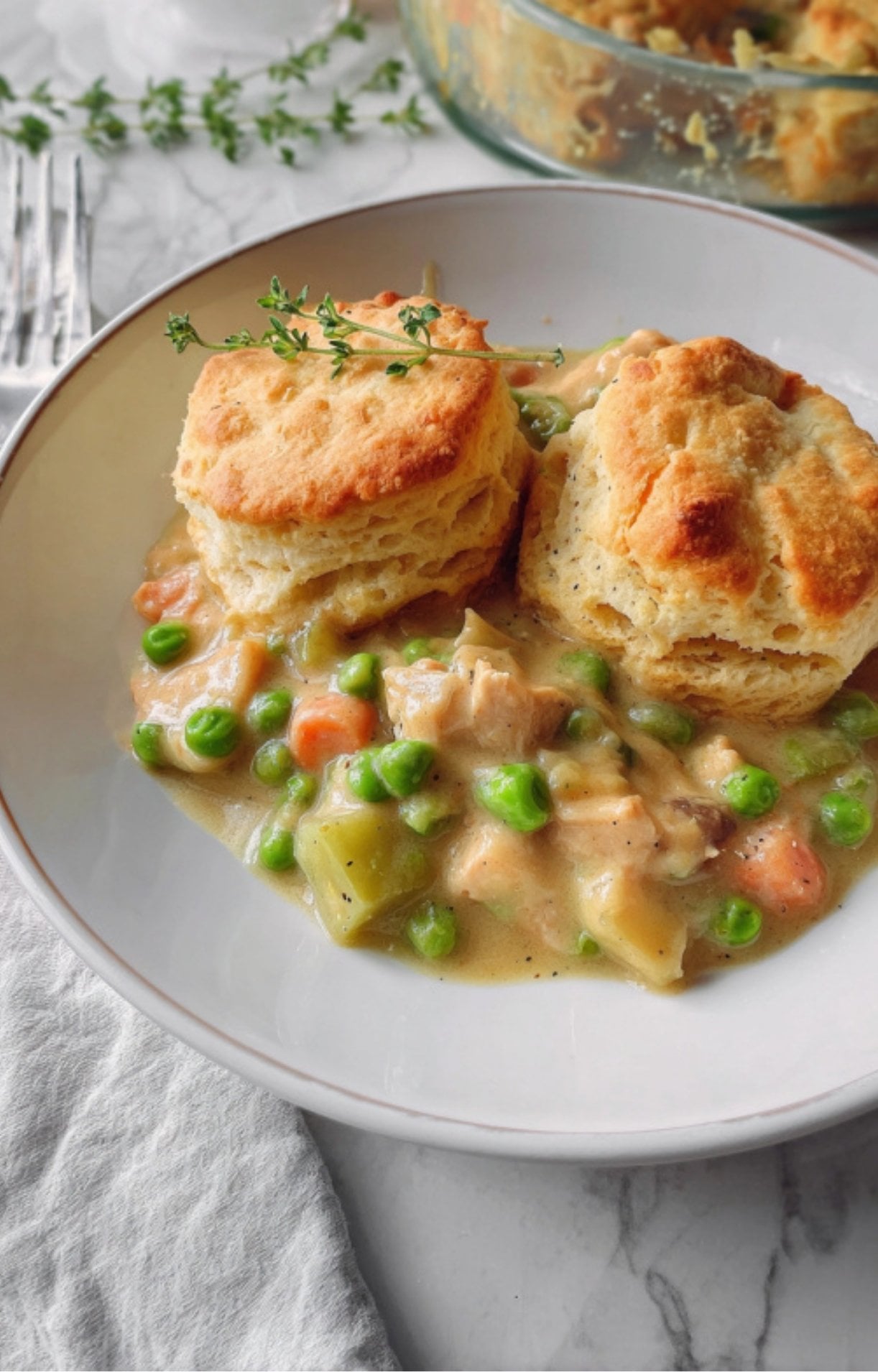 A white bowl filled with creamy turkey pot pie filling featuring peas and carrots, topped with two golden-brown flaky biscuits and a sprig of fresh thyme.