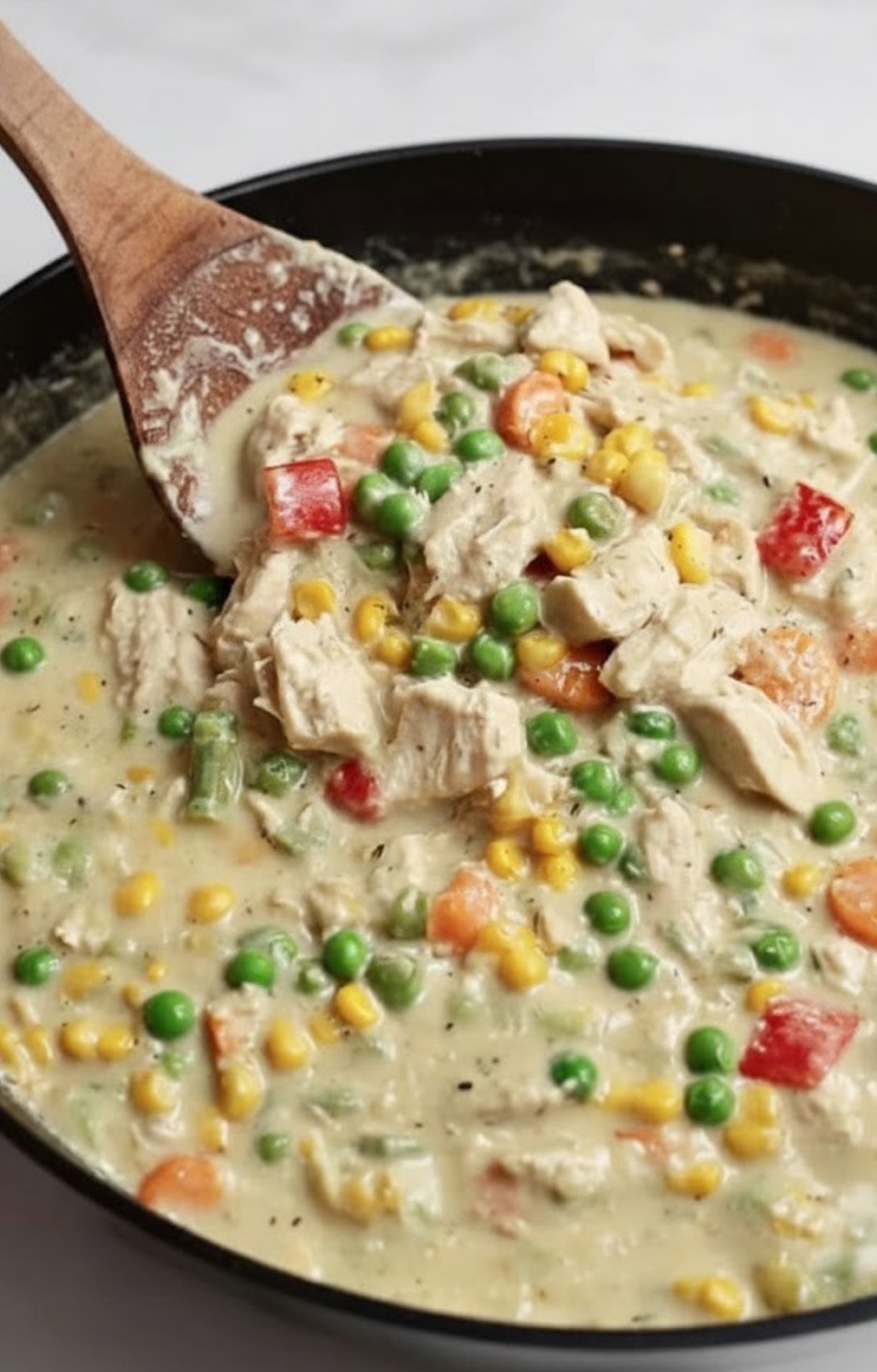 A wooden spoon stirring a thick, creamy mixture of shredded turkey, peas, corn, and carrots in a skillet for Turkey Pot Pie with Crescent Rolls.