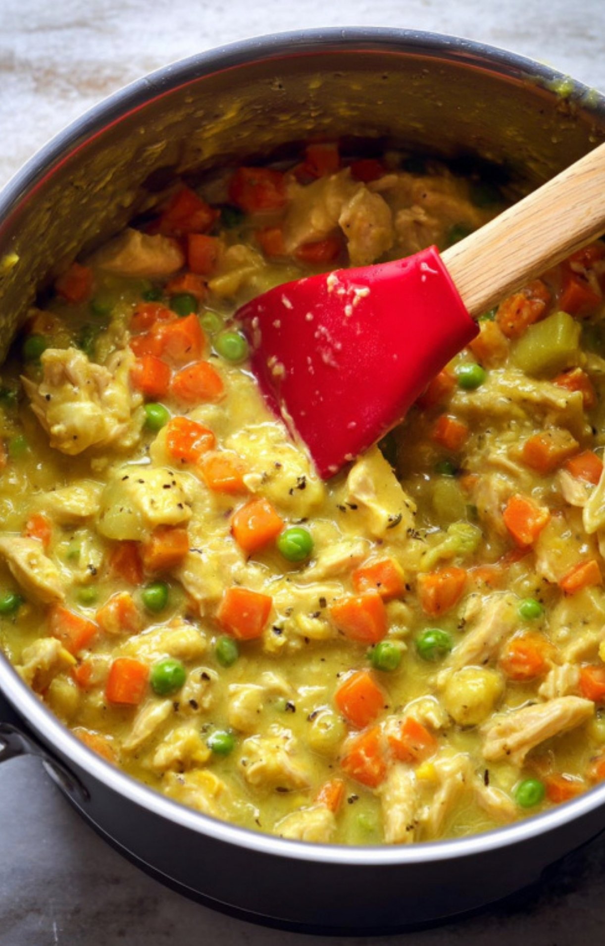 A close-up shot of a red spatula stirring a thick, creamy filling of turkey, peas, and carrots in a large stainless steel pot.