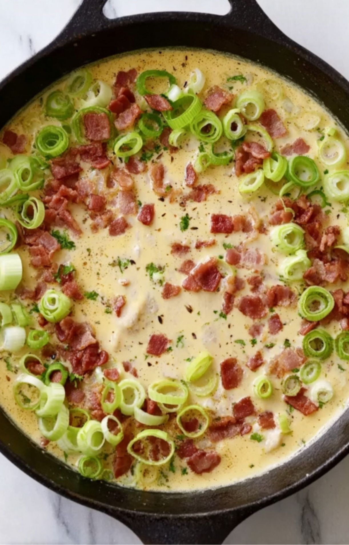 Sliced green leeks and crispy bacon pieces cooking in a creamy base within a black cast-iron skillet.
