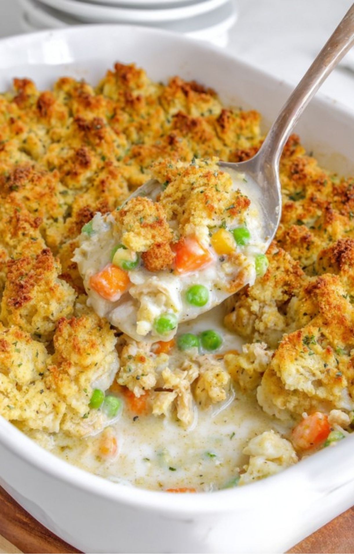A large metal spoon scooping out a serving of creamy turkey pot pie topped with golden-brown toasted stuffing from a white casserole dish.
