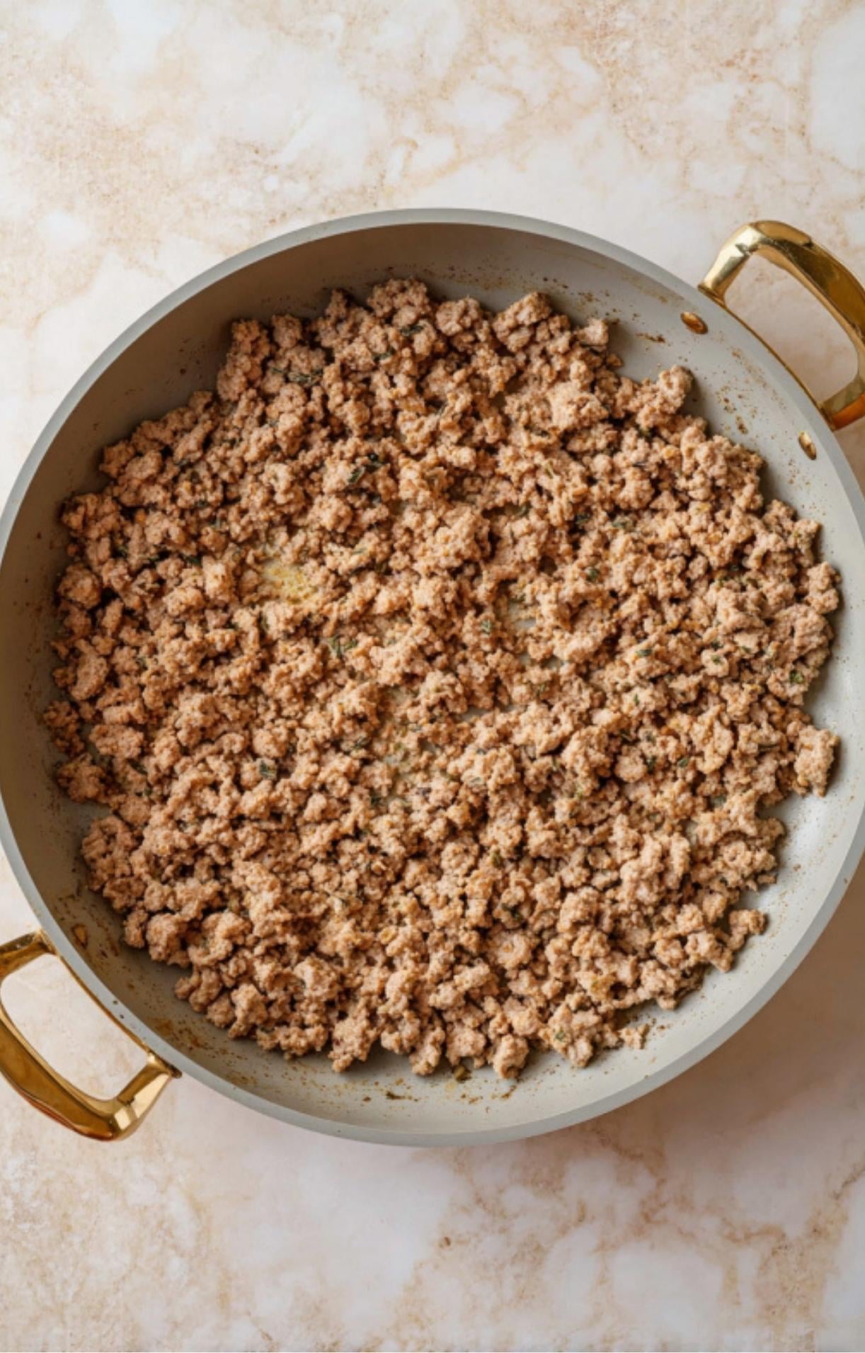 A large grey skillet with gold handles containing cooked and crumbled ground turkey being seared on a marble countertop.