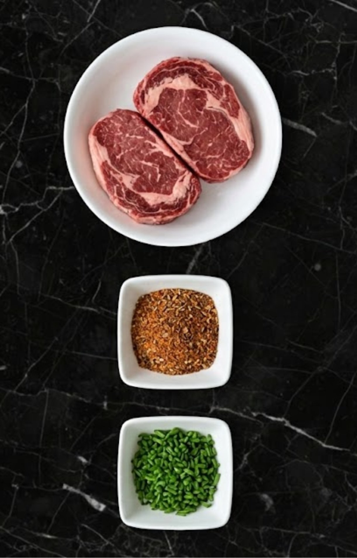 Two raw ribeye steaks in a white bowl alongside small dishes of dry rub seasoning and chopped fresh chives.