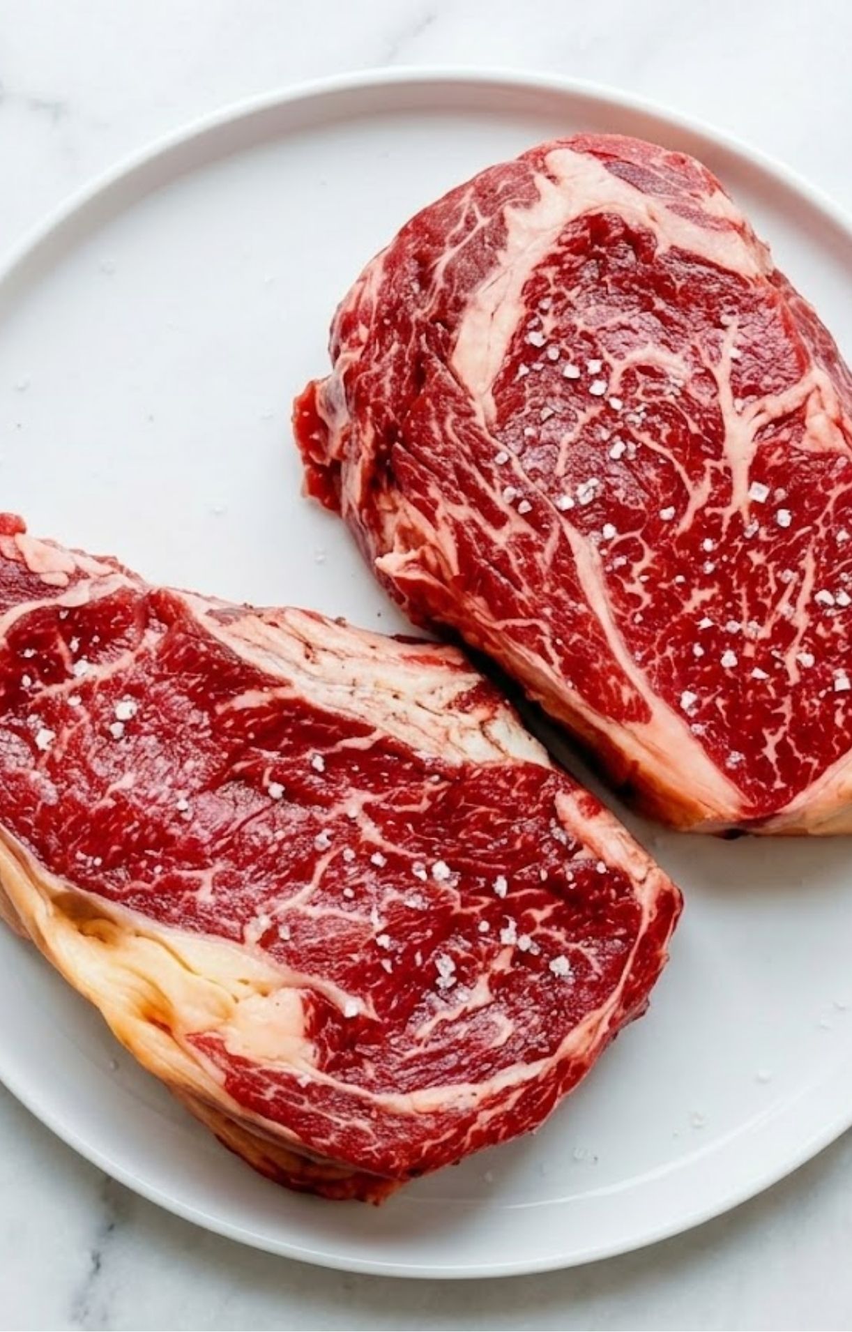 Two raw rib eye steaks on a white plate heavily seasoned with coarse sea salt crystals before cooking.