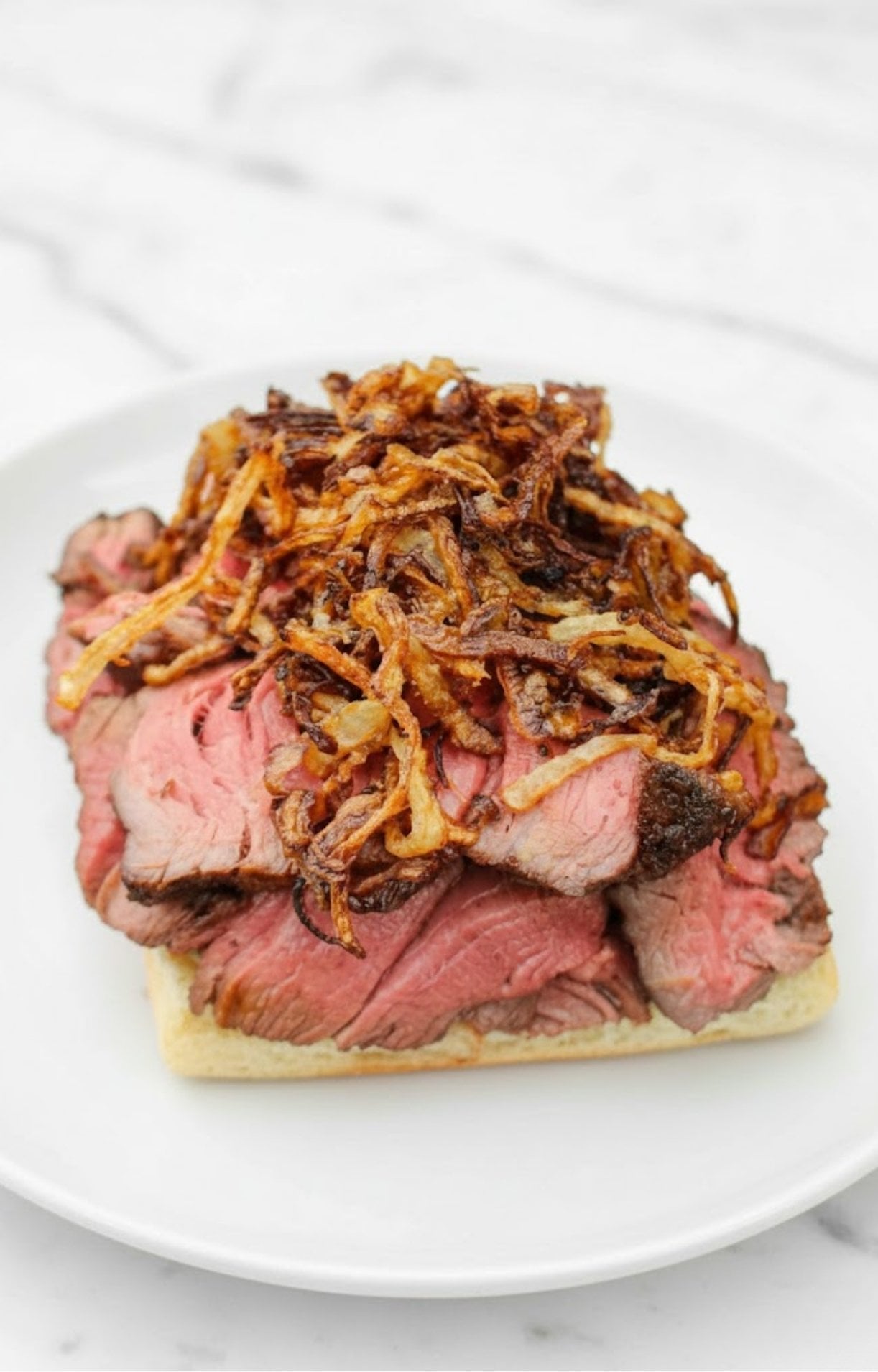 Sliced grilled steak piled high on toasted bread and topped with crispy fried onions, served on a white plate.