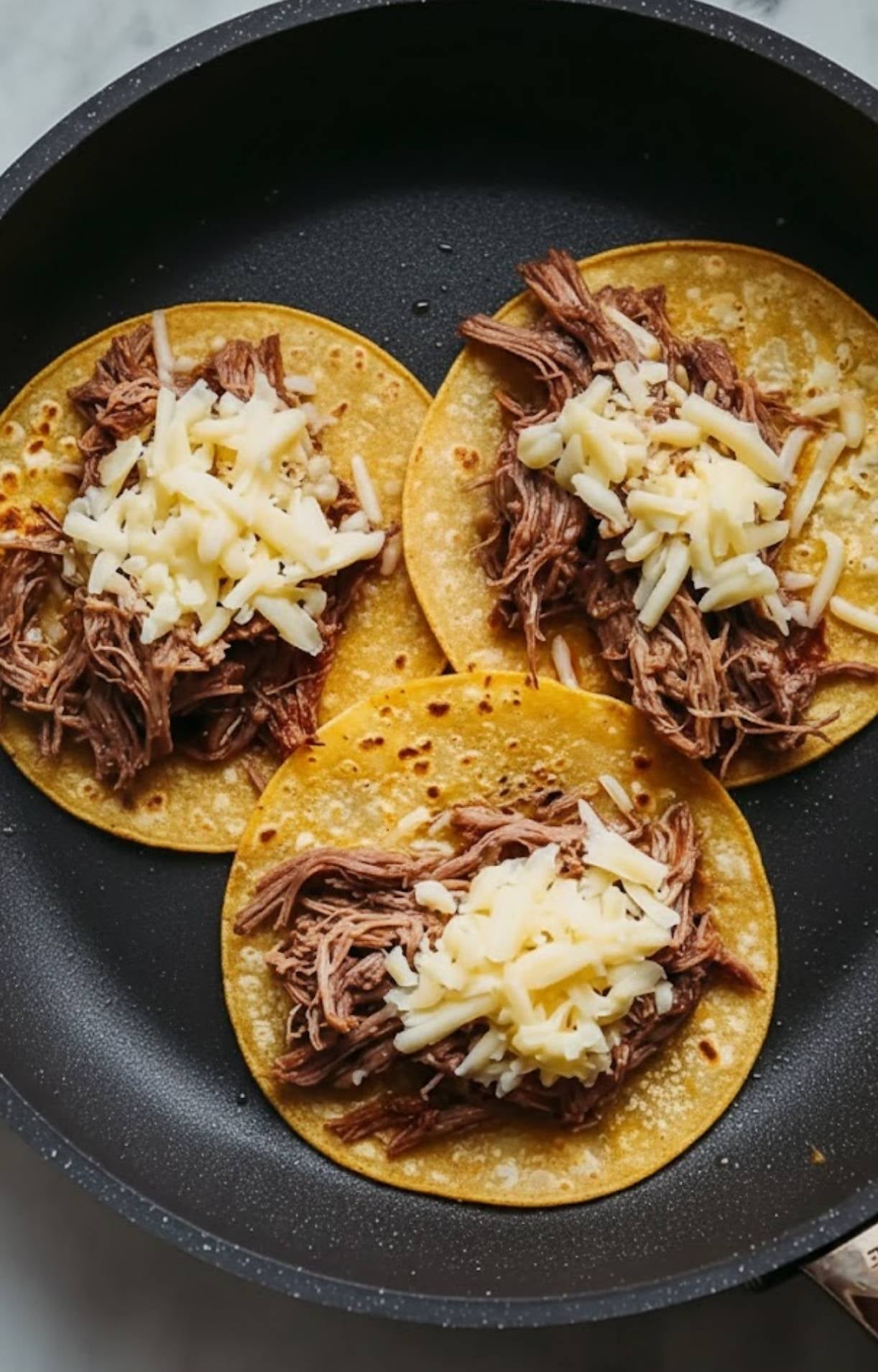 Three corn tortillas in a black skillet topped with generous portions of shredded beef and mounds of white cheese as they melt together.