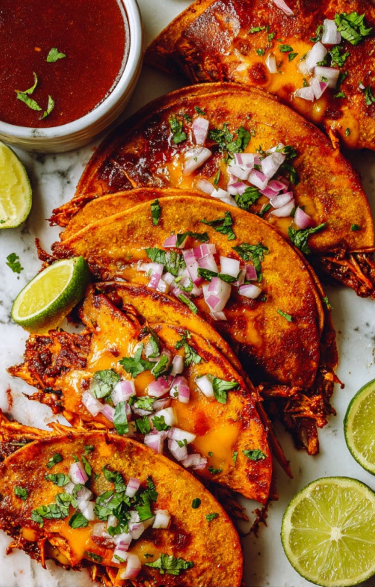 A row of golden-brown chicken birria tacos lined up on a marble surface, topped with a vibrant mix of onions and cilantro with lime wedges on the side.