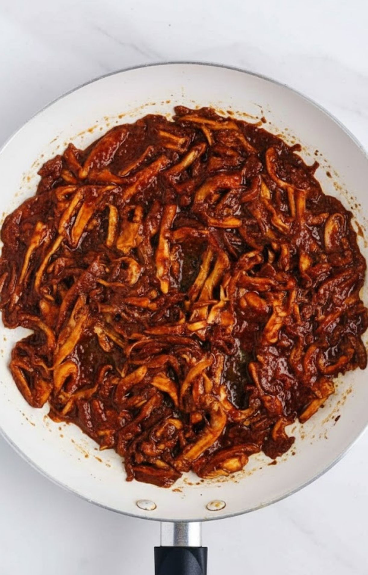 Shredded mushrooms thoroughly coated in a thick, dark red birria sauce in a white frying pan.