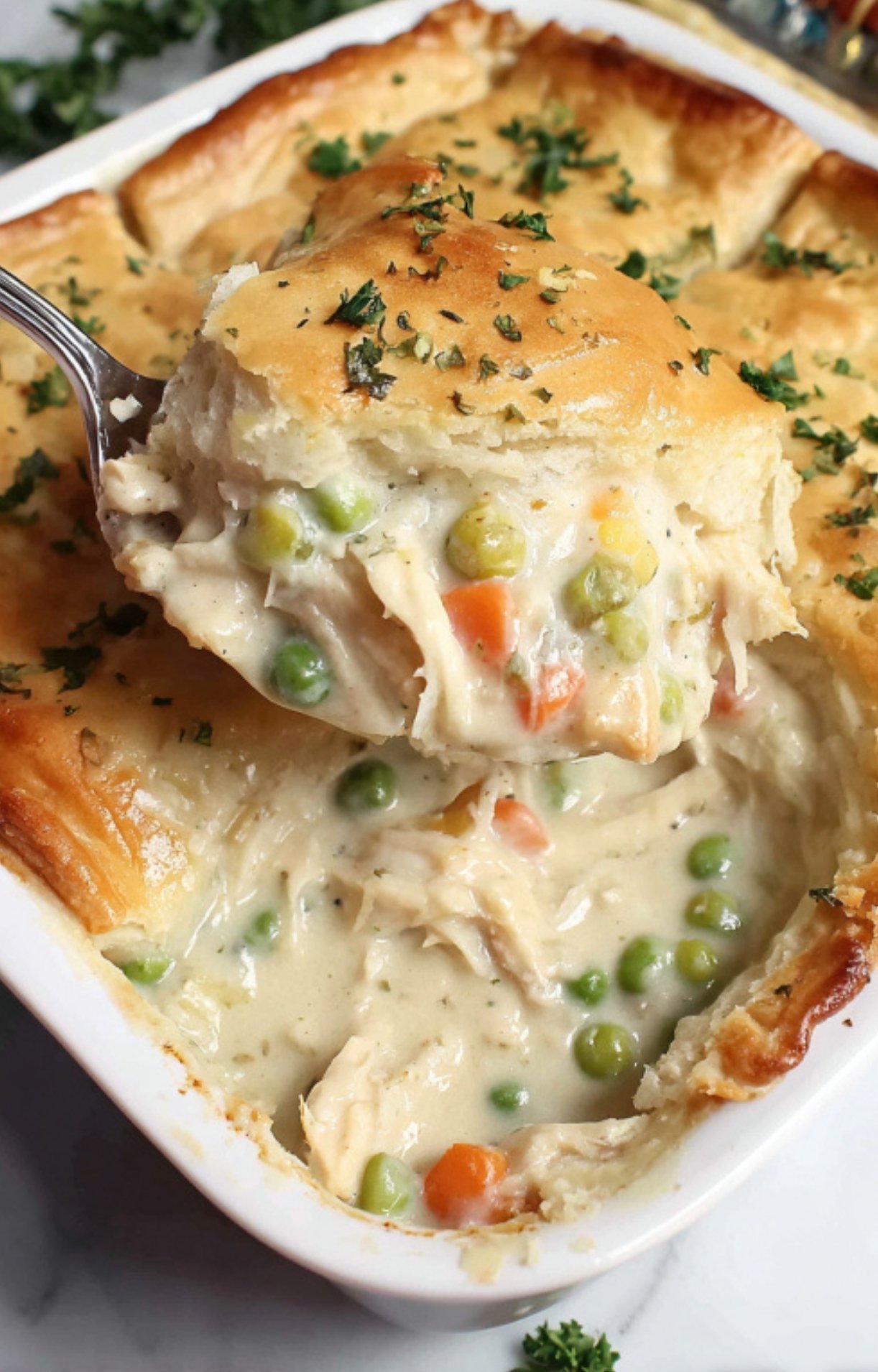 A large spoon scooping a portion of golden-brown pot pie out of the baking dish, garnished with fresh parsley.