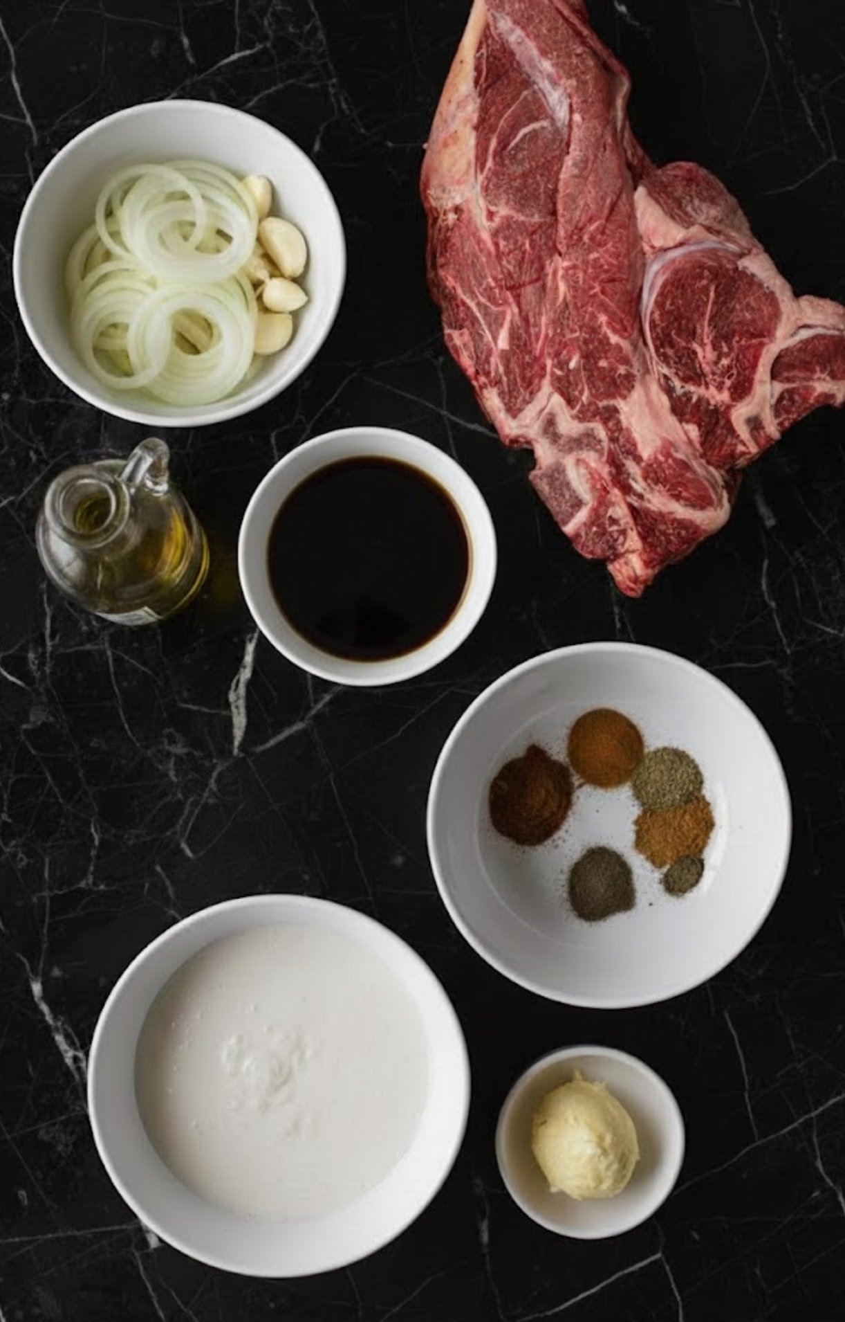 An overhead shot of raw chuck roast, sliced onions, garlic cloves, olive oil, beef broth, butter, and various spices in white bowls.