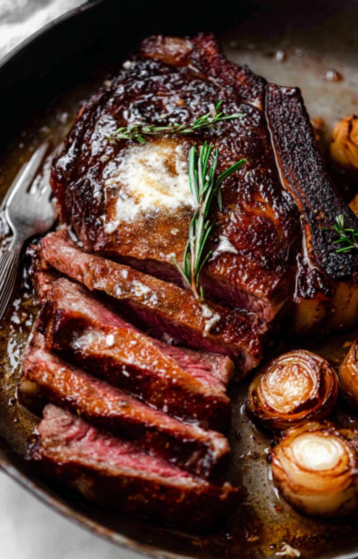 A close-up of a thick-cut dry aged rib eye steak in a cast iron skillet, topped with melting herb butter, fresh rosemary, and charred shallots.