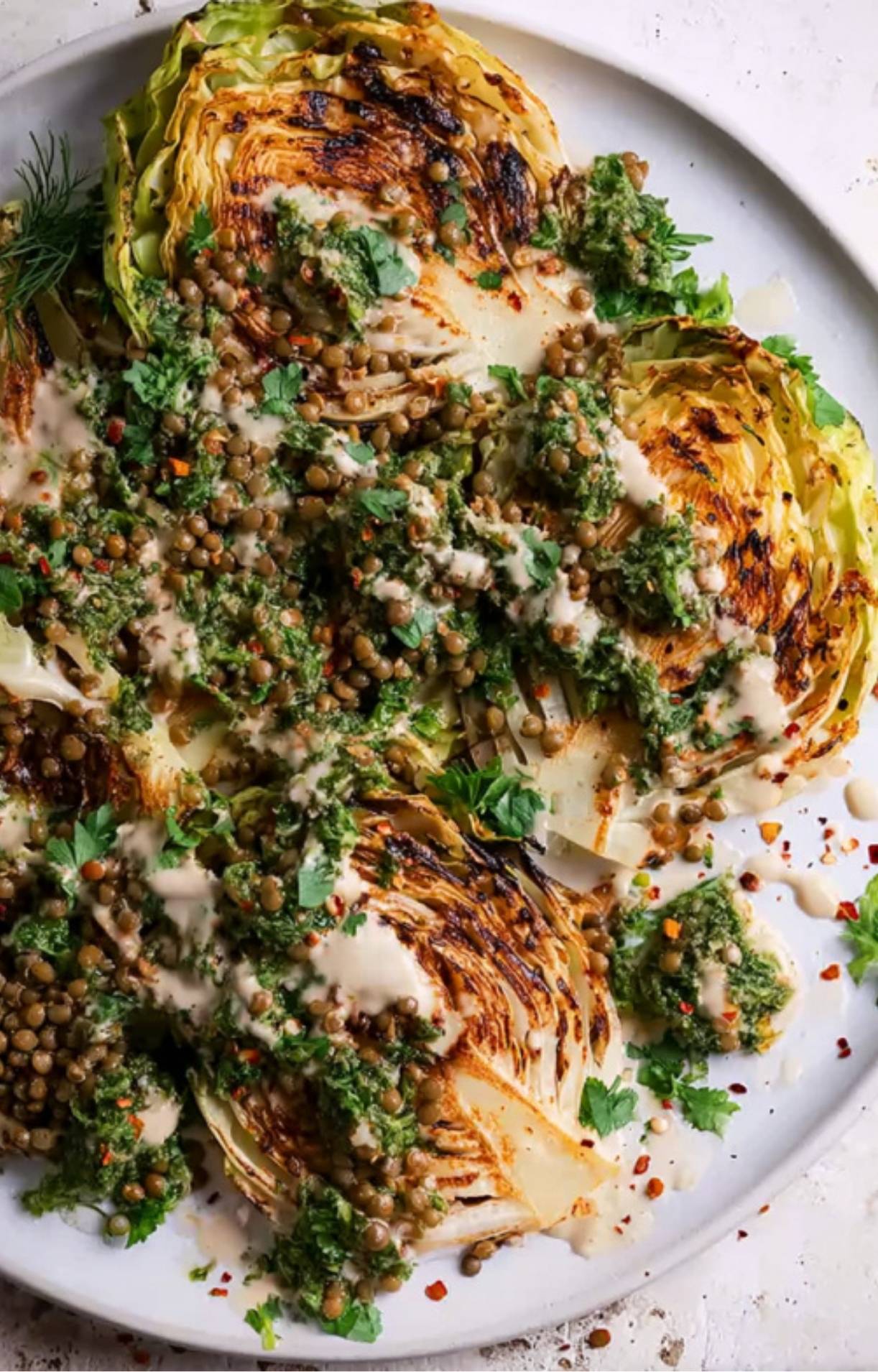 Large platter of grilled cabbage steaks topped with lentils, chimichurri, and a drizzle of tahini sauce.