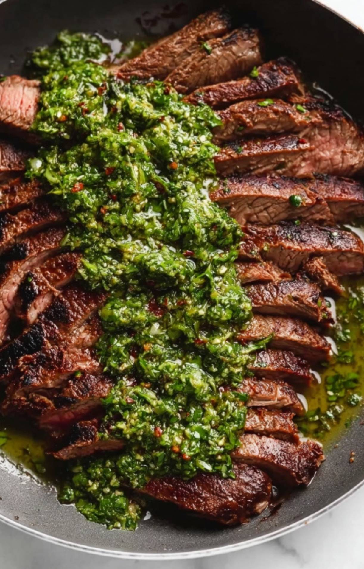Thinly sliced grilled flank steak resting in a dark skillet, topped with a thick layer of fresh chimichurri sauce showing bits of red chili and garlic.