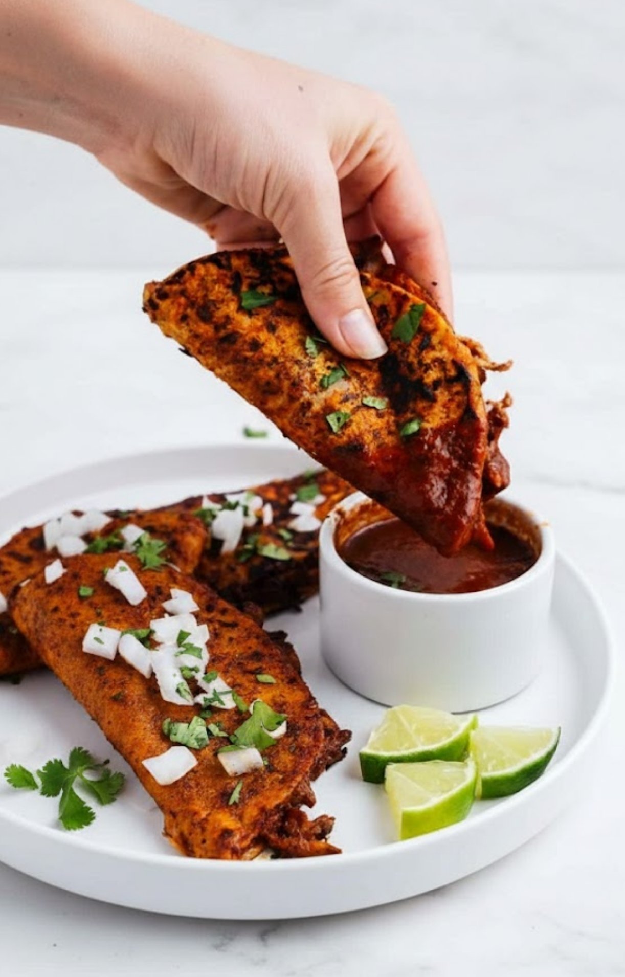 A hand holding a crispy, golden-brown vegan birria taco and dipping it into a small bowl of rich red consommé.