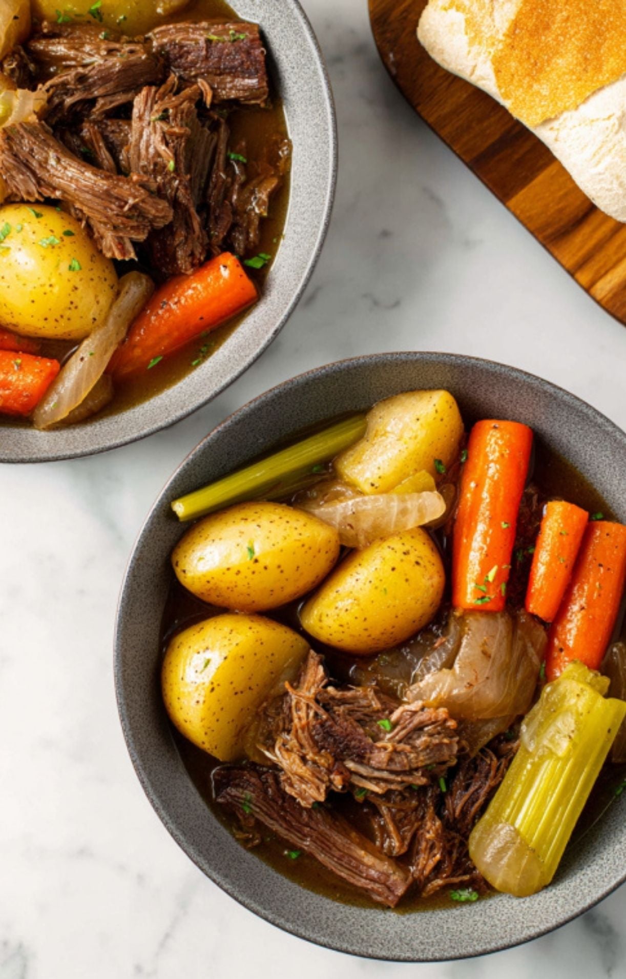 Two grey bowls served with hearty portions of pot roast, vegetables, and a side of crusty bread.