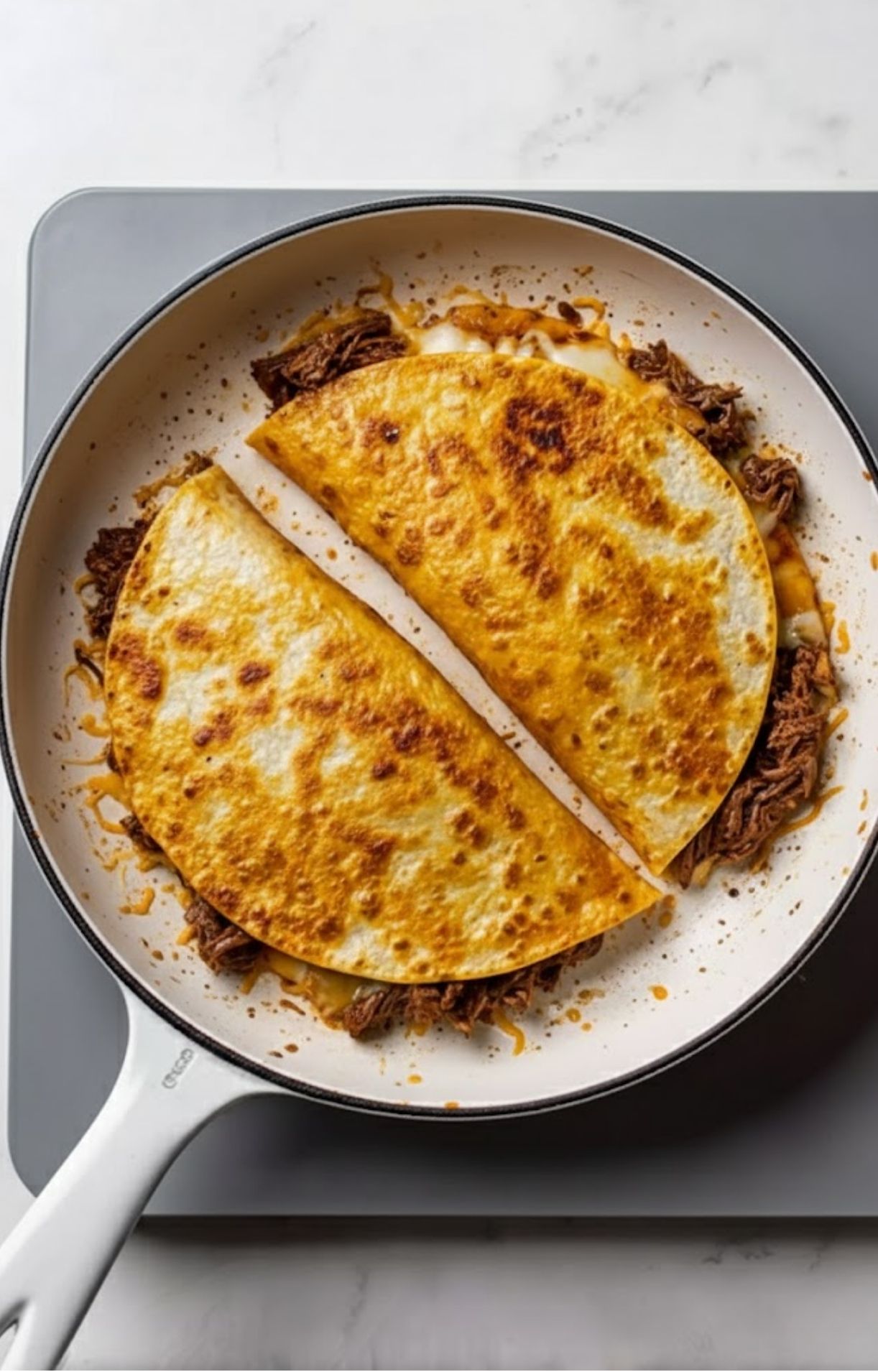 Two corn tortillas folded over shredded beef and melted cheese, crisping in a white skillet until golden brown.