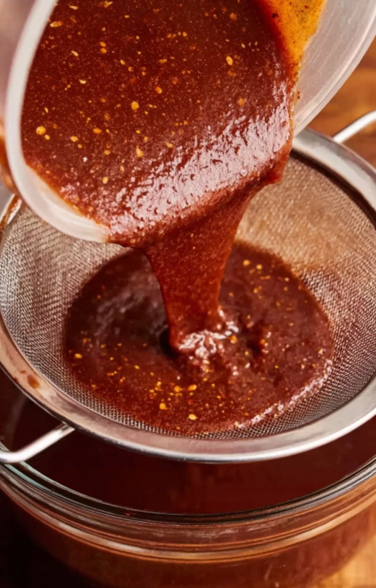 Thick red birria sauce being poured through a fine-mesh metal strainer into a glass bowl to create a smooth dipping consommé.
