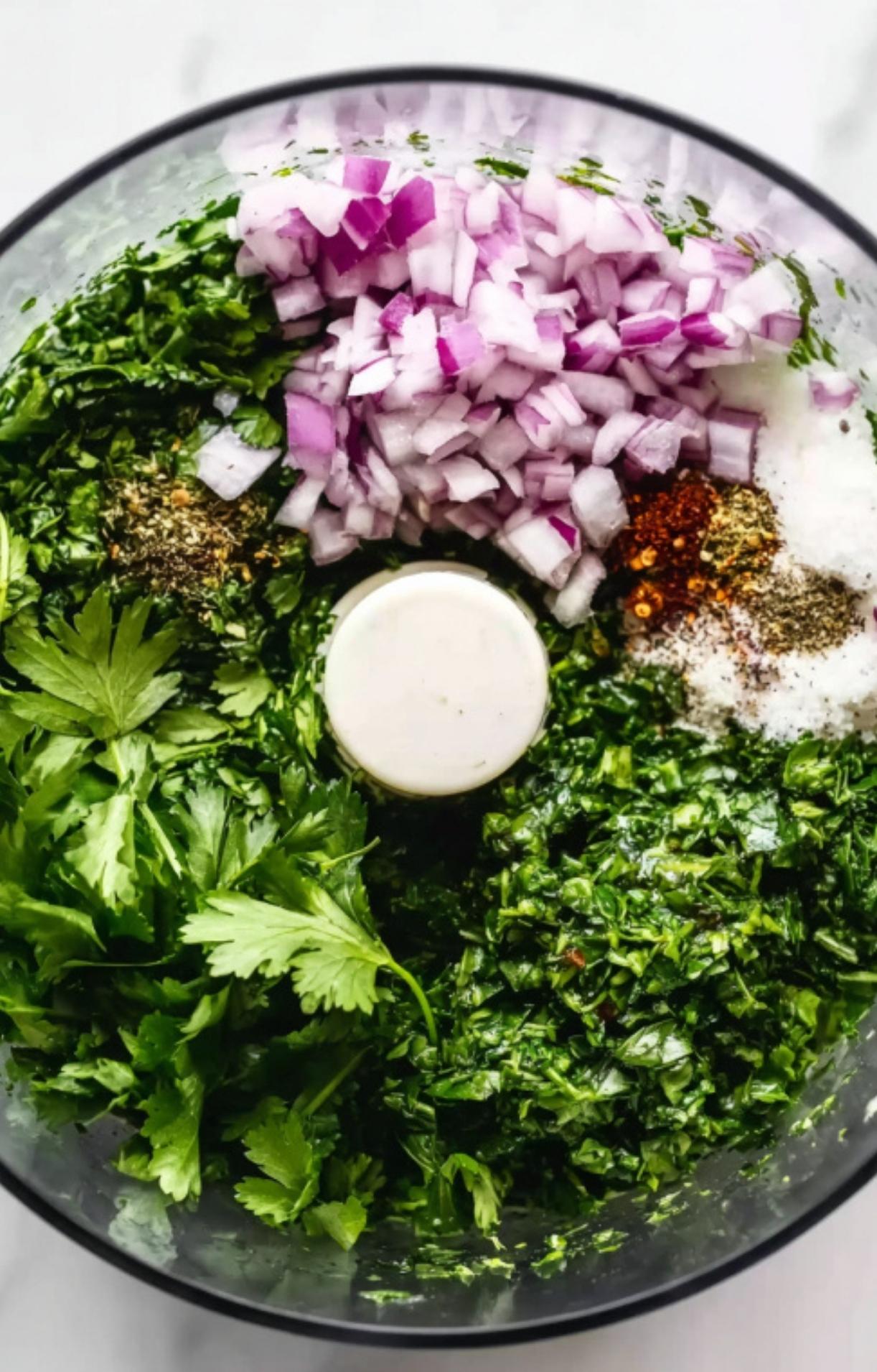 A food processor bowl containing fresh cilantro, parsley, diced red onion, garlic, and various dried spices ready to be blended into a sauce for Grilled Flank Steak.