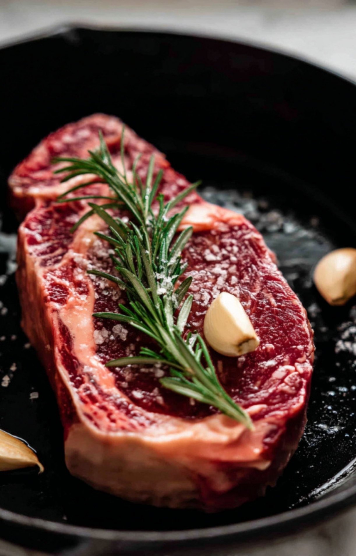 A raw dry aged rib eye steak in a black pan seasoned heavily with coarse sea salt, topped with a sprig of rosemary and garlic for aromatics.