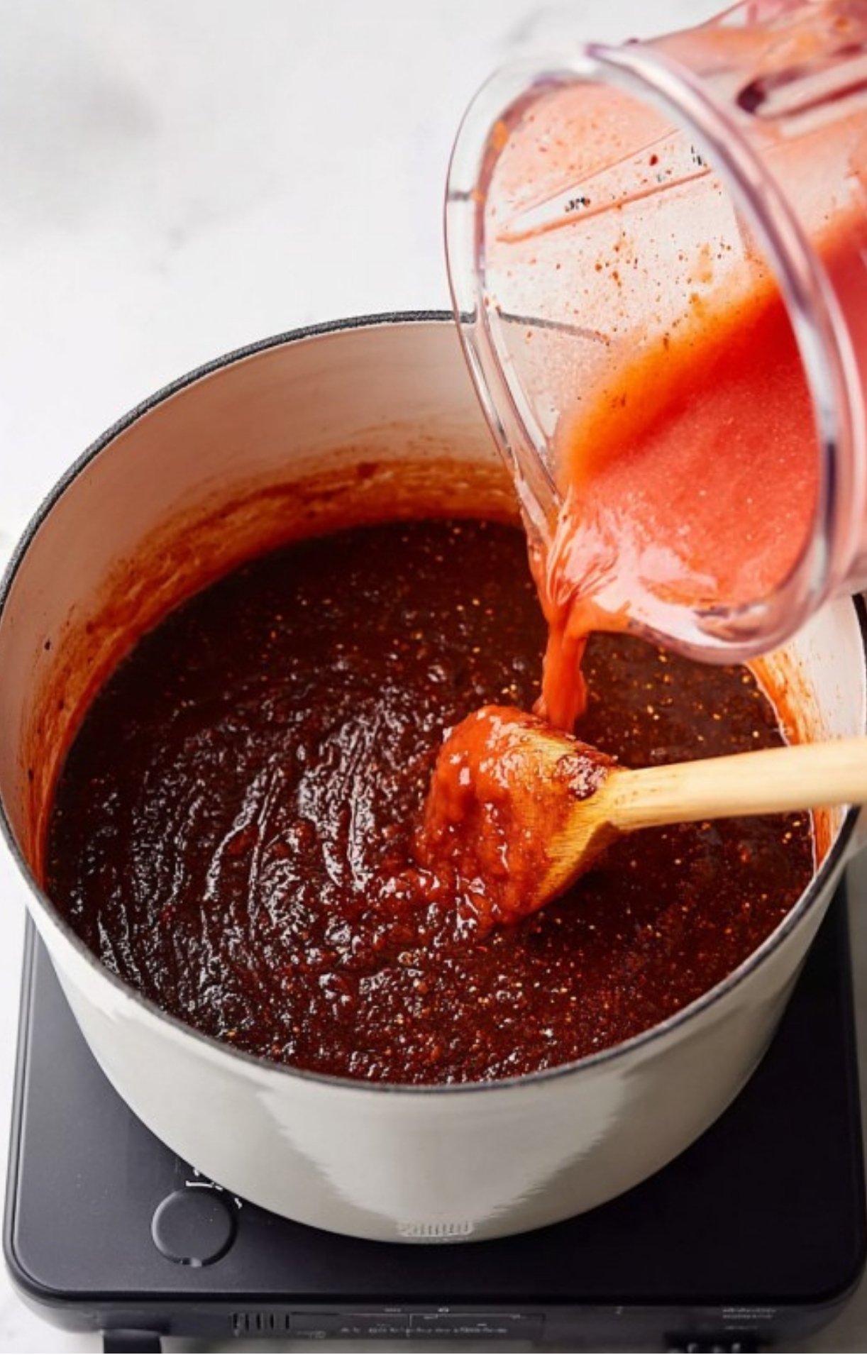 A vibrant red tomato and chili mixture being poured from a blender into the dark, simmering birria base.