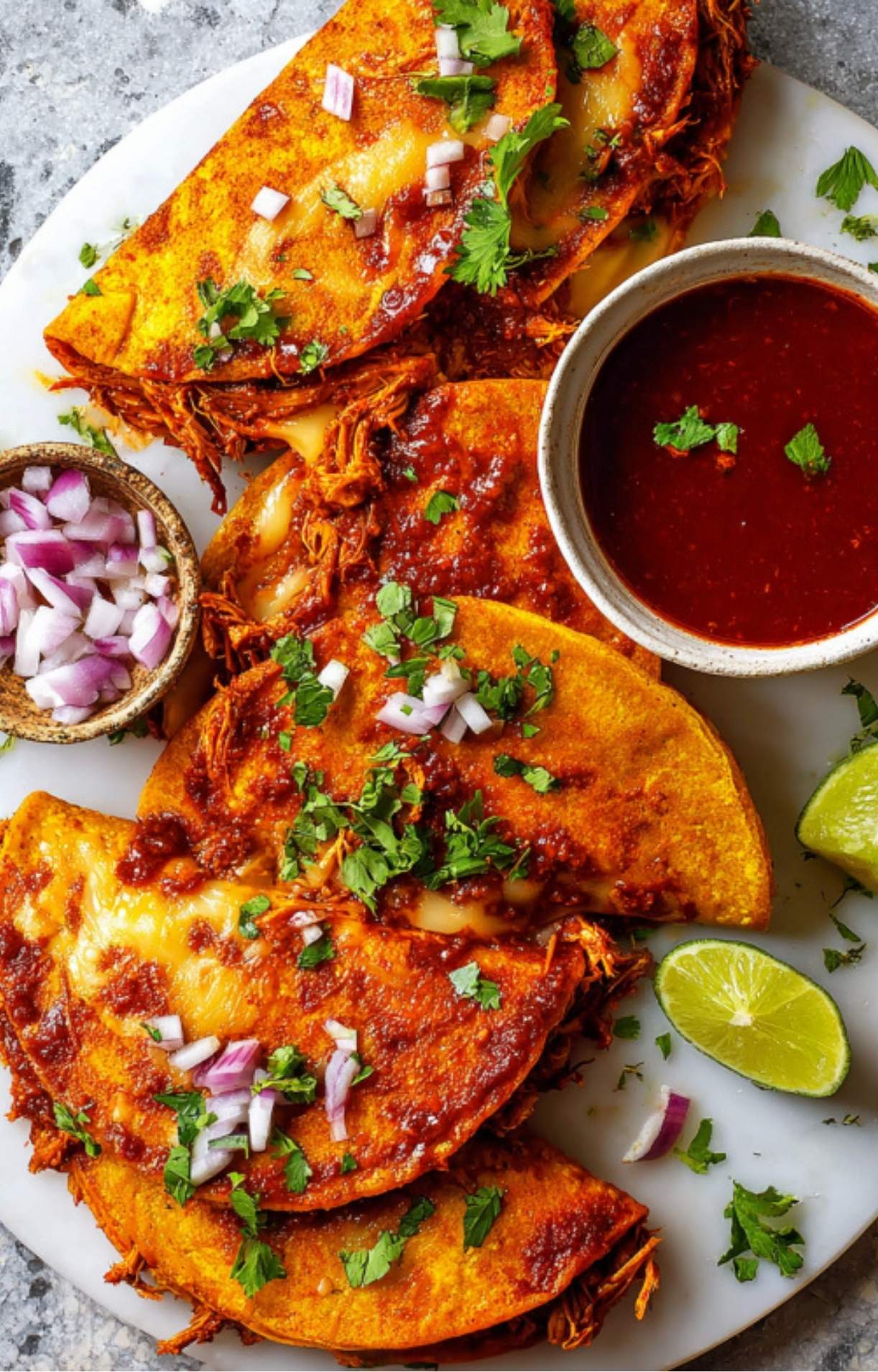An overhead view of a full platter of birria tacos, extra garnishes in small bowls, and the essential dipping sauce.
