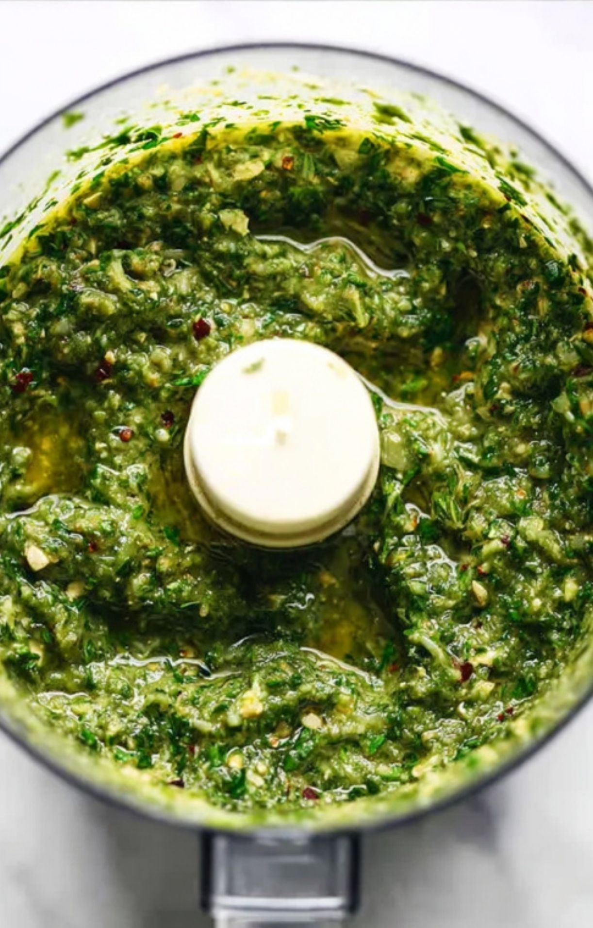 A food processor filled with a freshly pulsed green sauce, showing a coarse texture of chopped herbs, garlic, and oil.