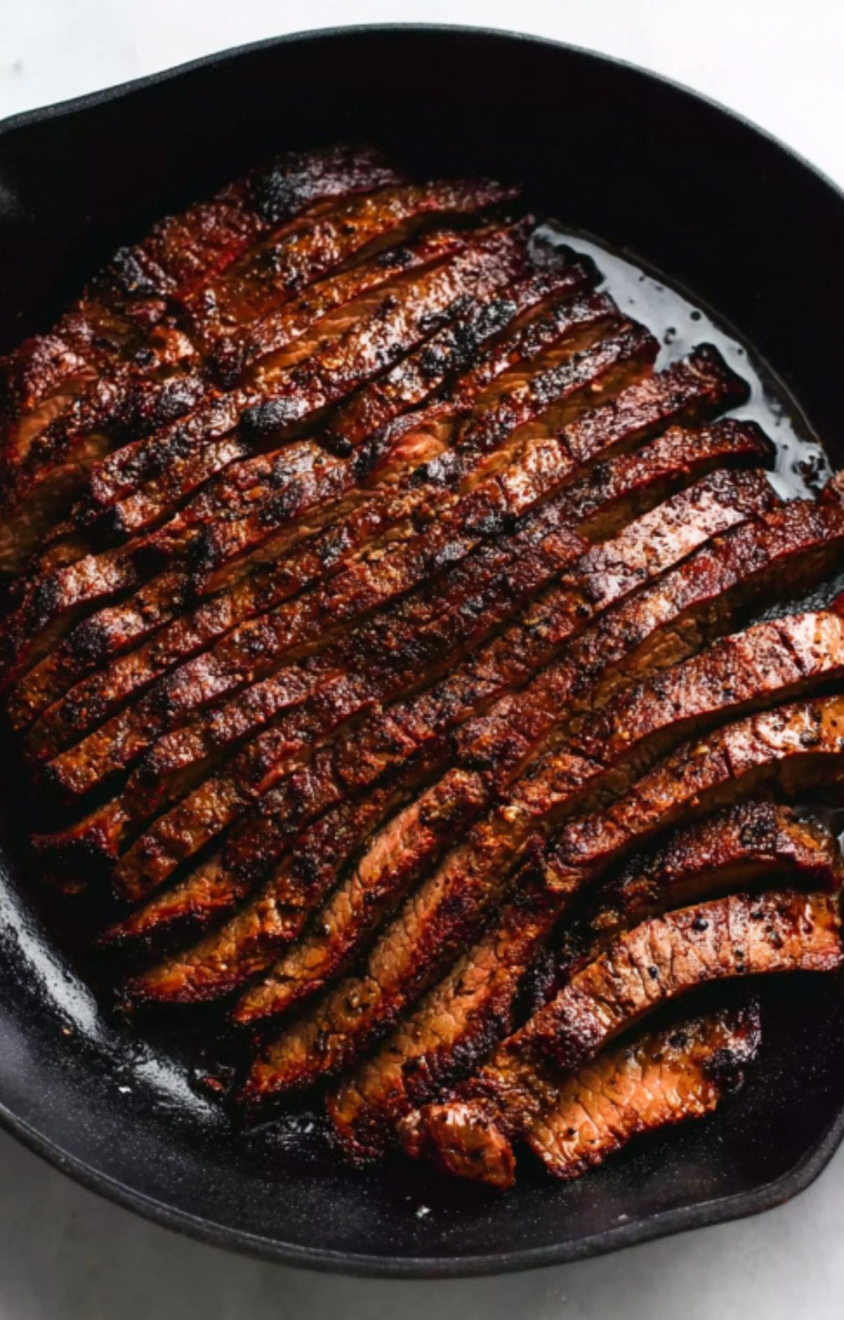 A close-up of a well-seared flank steak sliced against the grain and arranged neatly in a black cast-iron pan, glistening with natural juices.